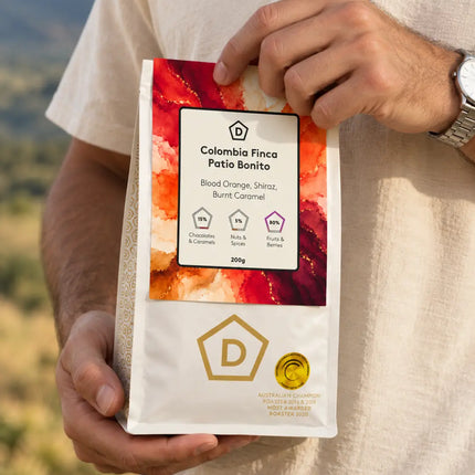A person outdoors holding a bag of Colombia Finca Patio Bonito coffee. The bag is white with a colorful red, orange, and yellow watercolor-style label and gold branding details.

